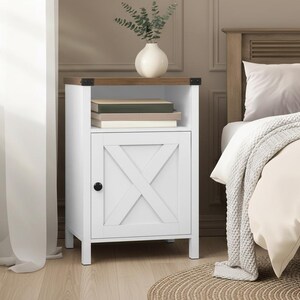 May include: White bedside cabinet with a wooden top and a door featuring an X-shaped design. The cabinet has a black knob and is next to a bed with a neutral-colored comforter. A vase with greenery sits on top.