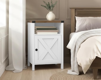 May include: White bedside cabinet with a wooden top and a door featuring an X-shaped design. The cabinet has a black knob and is next to a bed with a neutral-colored comforter. A vase with greenery sits on top.