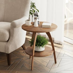 May include: A round, two-tiered wooden side table with a warm brown finish. The table has a small plant on the lower tier and a coffee cup, books, and a grinder on the top. A beige armchair is in the background.