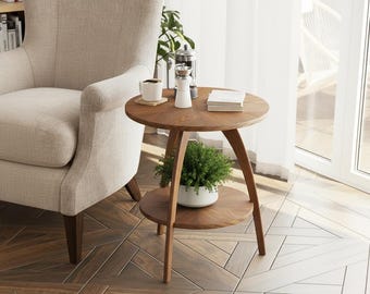 May include: A round, two-tiered wooden side table with a warm brown finish. The table has a small plant on the lower tier and a coffee cup, books, and a grinder on the top. A beige armchair is in the background.