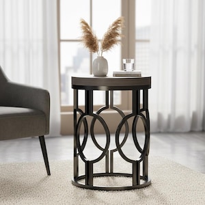 May include: A round side table with a dark metal base featuring an open, geometric design. The tabletop is a light wood color. A small white vase with dried plants, a glass, and a book are on top. A gray armchair is to the left.