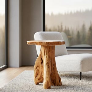 May include: A natural wood side table with a unique, organic design. The table has a round top and a textured, sculptural base. The light brown table is placed on a light-colored rug next to a white armchair.