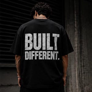 Könnte beinhalten: Schwarzes T-Shirt mit dem weißen Aufdruck "BUILT DIFFERENT" in verwitterter Schrift. Das Shirt hat einen klassischen Rundhalsausschnitt. Das Bild zeigt die Rückseite des Shirts.