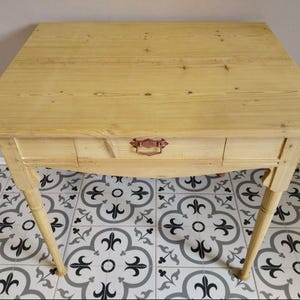 Può includere: Un tavolo in legno chiaro con un singolo cassetto e una maniglia decorativa. Il tavolo ha quattro gambe angolate e un piano quadrato. La venatura del legno è visibile. Il tavolo è largo circa 76 cm e lungo 76 cm.