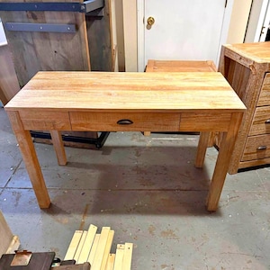 May include: A light-coloured wooden desk with a single drawer and a dark handle. The desk has four legs and a rectangular top. The wood grain is visible. A matching wooden cabinet is visible in the background.