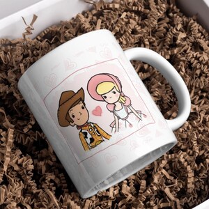 K&ouml;nnte beinhalten: Wei&szlig;e Keramik-Tasse mit einer Cartoon-Illustration von Woody und Bo Peep aus Toy Story. Das Design ist in einem rosa Rahmen mit Herzakzenten gehalten. Die Tasse hat einen wei&szlig;en Henkel und wird in einer Geschenkbox mit braunem Papier geliefert.