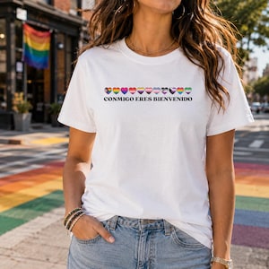 May include: A white short-sleeved t-shirt featuring a row of eleven small hearts, each filled with different pride flag color patterns. Below the hearts, the text CONMIGO ERES BIENVENIDO is printed in a bold, black, sans-serif typeface.