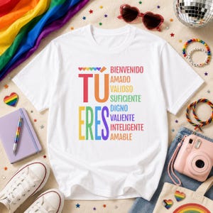 May include: A white short-sleeve t-shirt featuring the colorful text TU ERES BIENVENIDO AMADO VALIOSO SUFICIENTE DIGNO VALIENTE INTELIGENTE AMABLE in a rainbow-inspired palette, centered on the chest with a small row of rainbow hearts above.