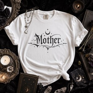 May include: White t-shirt with the word "Mother" in a gothic font, adorned with crescent moons and stars. The shirt is knotted at the bottom. The design is in black. The shirt is on a black surface with gothic-themed accessories.