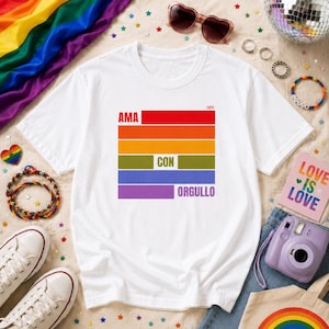 May include: A white short-sleeved t-shirt featuring a graphic of six horizontal stripes in red, orange, yellow, green, blue, and purple. The text "AMA" is printed in white on the red stripe, "CON" on the green stripe, and "ORGULLO" on the purple stripe.