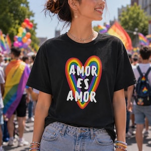 May include: A black short-sleeve t-shirt featuring a large rainbow-colored heart graphic on the chest. Inside the heart, the white text reads AMOR ES AMOR in a bold, sans-serif font. The shirt has a crew neckline and a standard fit.