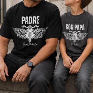 May include: A black t-shirt with the white text PADRE and a graphic of a motorcycle with wings, plus the text Guía y Protector. Beside it, a charcoal gray t-shirt with the white text CON PAPÁ, a matching motorcycle graphic, and the text Voy Seguro.