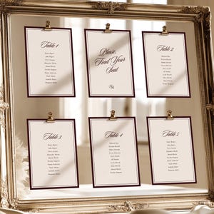 Puede incluir: Un marco de fotos grande y ornamentado en tono dorado que sostiene seis tarjetas de color crema con bordes rojo oscuro. Las tarjetas están suspendidas por clips dorados y presentan texto negro, incluyendo "Please, Find Your Seat".
