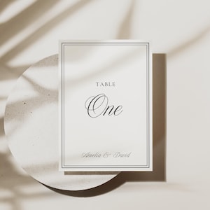 Puede incluir: Una tarjeta de número de mesa color crema con un borde negro. La tarjeta dice "TABLE One" en una elegante escritura, con "Amelia & David" debajo. La tarjeta está sobre una superficie circular texturizada.