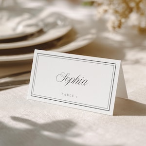 May include: A white place card with the name "Sophia" in elegant script, framed by a thin black border. The text "TABLE 1" is below the name. The card is folded and stands on a textured surface, suggesting a wedding or event setting.