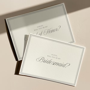 May include: Two ivory invitation cards with black text and a black border. One card reads "Sarah, Will you be my Maid of Honour?" and the other reads "Olivia, Will you be my Bridesmaid?" The cards are on a light beige surface.