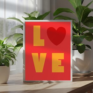 Puede incluir: Una impresión artística enmarcada con la palabra "LOVE" en letras amarillas y un corazón rojo sobre un fondo rojo. La obra de arte se exhibe sobre una superficie de madera, con plantas verdes en macetas blancas en el fondo.