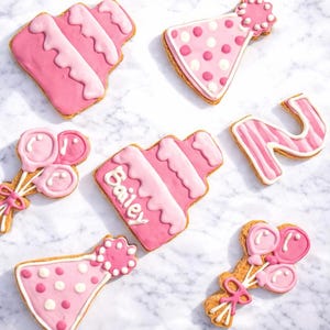 May include: Pink frosted cookies in various shapes, including a cake, party hat, balloons, and the number 2. One cake cookie has the name "Bailey" written on it. The cookies are decorated with white and pink icing.