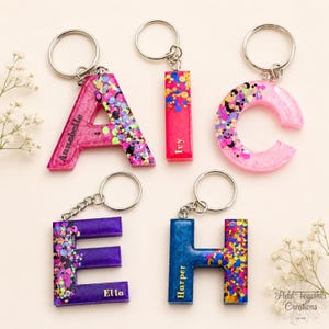 May include: Resin keyrings in the shape of the letters A, C, E, H, and I. Each letter is a different colour, with glitter and a name. The keyrings are attached to silver key rings.