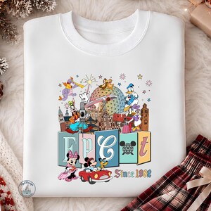 May include: A white crewneck sweatshirt featuring a colorful graphic print of Disney characters including Mickey, Minnie, Donald, Daisy, Goofy, Pluto, and Figment. The design includes the text EPCOT and Since 1982, with icons of the park landmarks.
