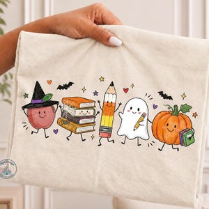 May include: A cream-colored fabric item featuring a colorful cartoon print of a smiling apple wearing a witch hat, a stack of books with a spider web, a pencil, a ghost holding a pencil, and a pumpkin holding a book, accented by bats and stars.