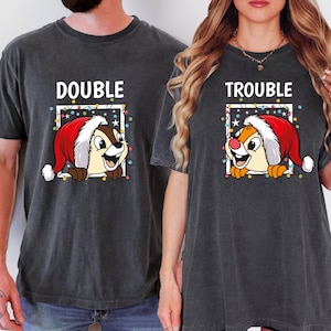 May include: Two charcoal gray t-shirts featuring a graphic of a cartoon chipmunk wearing a red Santa hat. The left shirt has the text DOUBLE above the chipmunk, and the right shirt has the text TROUBLE above the chipmunk, both framed by Christmas lights.