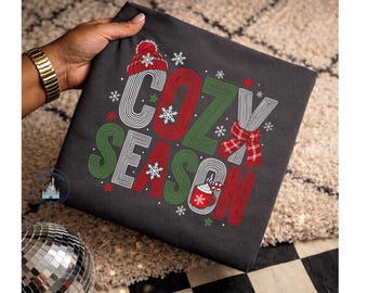 May include: A dark gray t-shirt featuring the text COZY SEASON in red, green, and white striped lettering. The design includes a red knit hat, a red plaid scarf, a red mug with a candy cane, and white snowflakes scattered throughout.