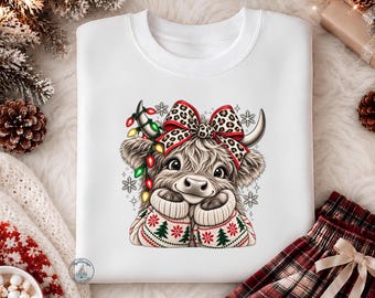 May include: A white crewneck sweatshirt featuring a graphic of a fluffy highland cow wearing a patterned sweater with Christmas trees and snowflakes. The cow has a leopard print bow on its head and is wrapped in colorful Christmas light bulbs.