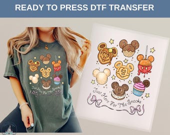 Può includere: Una t-shirt verde scuro con una stampa grafica di vari snack a forma di orecchie di topo, tra cui un waffle, un pretzel, un cupcake e una mela caramellata, con il testo Just Here For The Snacks e un disegno a nastro sotto gli snack.