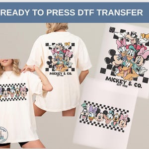 May include: Cream-colored t-shirts featuring a cartoon graphic of Mickey Mouse and friends, with a black and white checkered border. The text "MICKEY & CO. EST 1928" is also visible. The image also shows a DTF transfer.