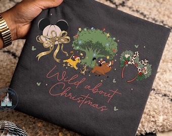 May include: A dark gray t-shirt featuring a graphic print with the text Wild about Christmas in pink script. The design includes a pair of mouse ears with a gold bow, a green tree with colorful lights, Simba, Timon, Pumbaa, and leopard print ears.