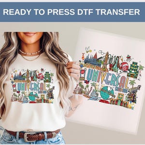 May include: A cream-colored t-shirt featuring a colorful graphic of Universal Studios attractions. The design includes the text "UNIVERSAL STUDIOS" with various illustrations of rides, landmarks, and characters. The words "READY TO PRESS DTF TRANSFER" are at the top.