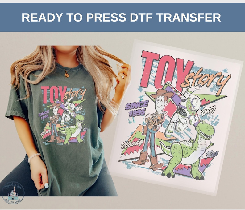 May include: A dark green t-shirt featuring a colorful graphic of Toy Story characters, including Woody, Buzz Lightyear, and Rex. The design includes the text "TOY STORY" and "SINCE 1995". The image also shows a DTF transfer.