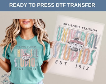 May include: A teal t-shirt with a colorful Universal Studios graphic, featuring the text "Orlando Florida" and "Est. 1912". A matching DTF transfer is held next to the shirt. The design includes pastel colors.