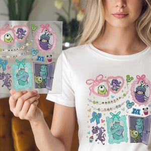 May include: A white t-shirt featuring a colorful graphic design of characters from the animated film "Monsters, Inc." The design includes images of Mike Wazowski, Sulley, and Boo, along with the text "Monsters, Inc."