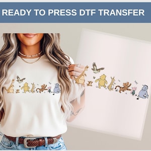May include: A cream-colored t-shirt featuring a line of cartoon characters, including a kangaroo, bear, rabbit, tiger, and pig. The text "READY TO PRESS DTF TRANSFER" is at the top. The design is on a white background.
