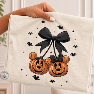 May include: A cream-colored t-shirt featuring a graphic of two orange pumpkin jack-o'-lanterns with mouse-shaped ears, connected by stems to a large black bow. The design includes small black bat silhouettes and star shapes scattered around.