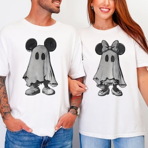 May include: Two white t-shirts featuring graphic prints of cartoon characters dressed as ghosts. The left shirt displays a character with round ears under a sheet, while the right shirt shows a character with a bow and ears under a sheet.