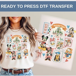 May include: A cream-colored t-shirt featuring a colorful Disney Trip design with Mickey Mouse, Minnie Mouse, Donald Duck, and other characters. The design includes the text "DISNEY TRIP 2026" and "LET'S GO! TRIP AWAITS".