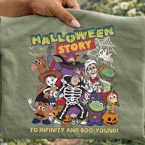 May include: An olive green t-shirt featuring a graphic of Toy Story characters in Halloween costumes, including a skeleton Buzz Lightyear, with the text HALLOWEEN STORY and TO INFINITY AND BOO YOUND printed in yellow and white lettering.