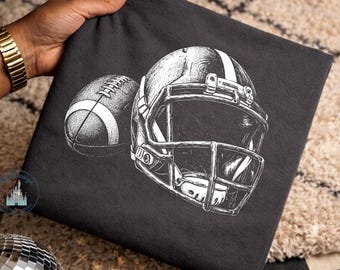 May include: A charcoal gray t-shirt featuring a white graphic illustration of an American football helmet and a football. The design uses detailed line work to depict the texture of the ball and the structure of the protective face mask.
