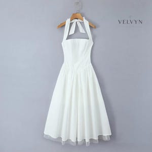 May include: A white halter dress with a flared skirt and a sheer hemline. The dress is hanging on a wooden hanger against a gray background. The dress has a bow at the neck and a fitted bodice. The brand name "VELVYN NOIR" is visible in the upper right corner.