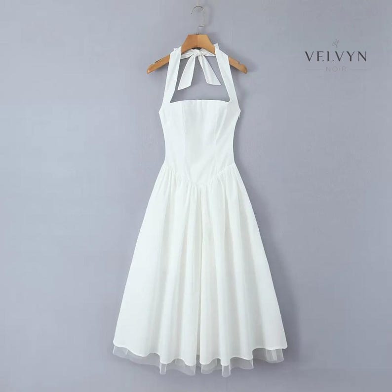 May include: A white halter dress with a flared skirt and a sheer hemline. The dress is hanging on a wooden hanger against a gray background. The dress has a bow at the neck and a fitted bodice. The brand name "VELVYN NOIR" is visible in the upper right corner.