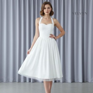 May include: A white halter dress with a sweetheart neckline and a flared skirt. The dress has a fitted bodice and a knee-length hemline. The skirt has a layer of tulle at the hem. The dress is worn by a person in front of a gray curtain.