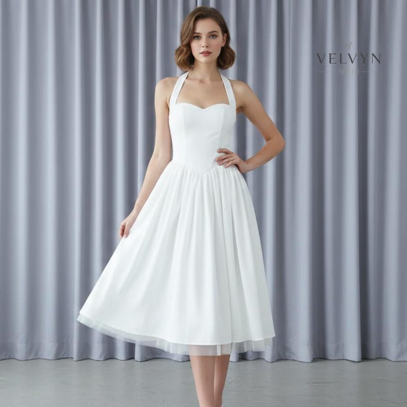 May include: A white halter dress with a sweetheart neckline and a flared skirt. The dress has a fitted bodice and a knee-length hemline. The skirt has a layer of tulle at the hem. The dress is worn by a person in front of a gray curtain.