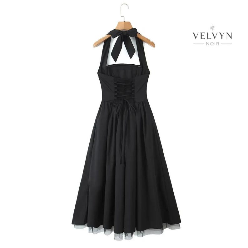 May include: Black halter dress with a bow at the neck and a corset-style back. The dress has a full skirt and is hanging on a wooden hanger. The brand name "VELVYN NOIR" is printed on the right side of the image.