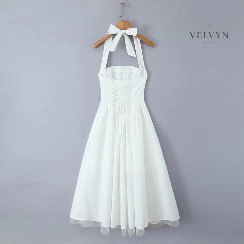 May include: A white halter dress with a bow detail at the neck, hanging on a wooden hanger. The dress features a fitted bodice with a lace-up back and a flowing skirt. The hemline is finished with a delicate trim.