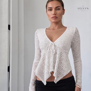 Puede incluir: Una blusa blanca de manga larga hecha de encaje transparente con escote en V y cierre de lazo frontal. La prenda presenta un dobladillo puntiagudo y asimétrico y mangas largas. El texto VELVYN NOIR está impreso en la esquina superior derecha.