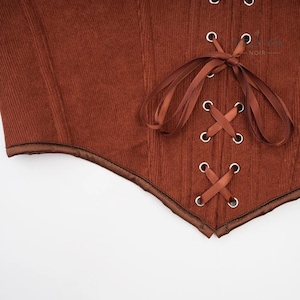 K&ouml;nnte beinhalten: Nahaufnahme eines braunen Korsetts mit Cordstruktur. Das Korsett hat eine Satinborte und ein Schn&uuml;rdesign mit silbernen &Ouml;sen. Das Gesamtdesign ist ein klassischer Vintage-Stil.