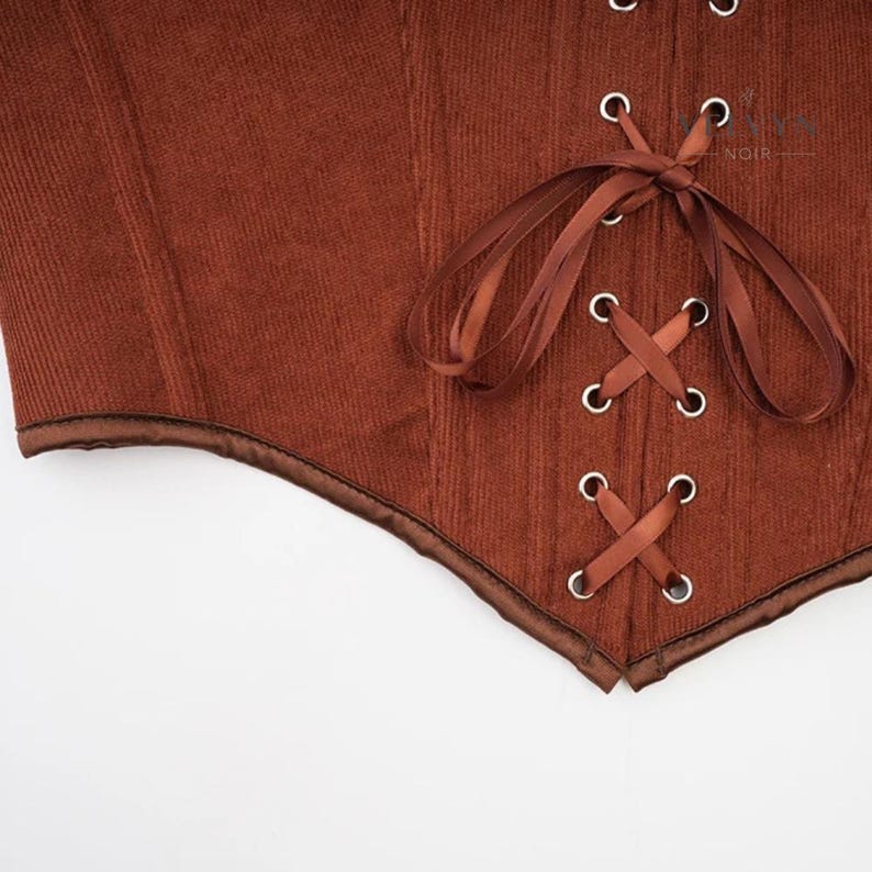 K&ouml;nnte beinhalten: Nahaufnahme eines braunen Korsetts mit Cordstruktur. Das Korsett hat eine Satinborte und ein Schn&uuml;rdesign mit silbernen &Ouml;sen. Das Gesamtdesign ist ein klassischer Vintage-Stil.